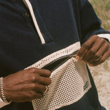 A close-up of a person outdoors wearing a navy long-sleeve pullover with white contrast trim and a large white mesh front pocket, with the zipper being opened. Sand and ocean are visible in the background.