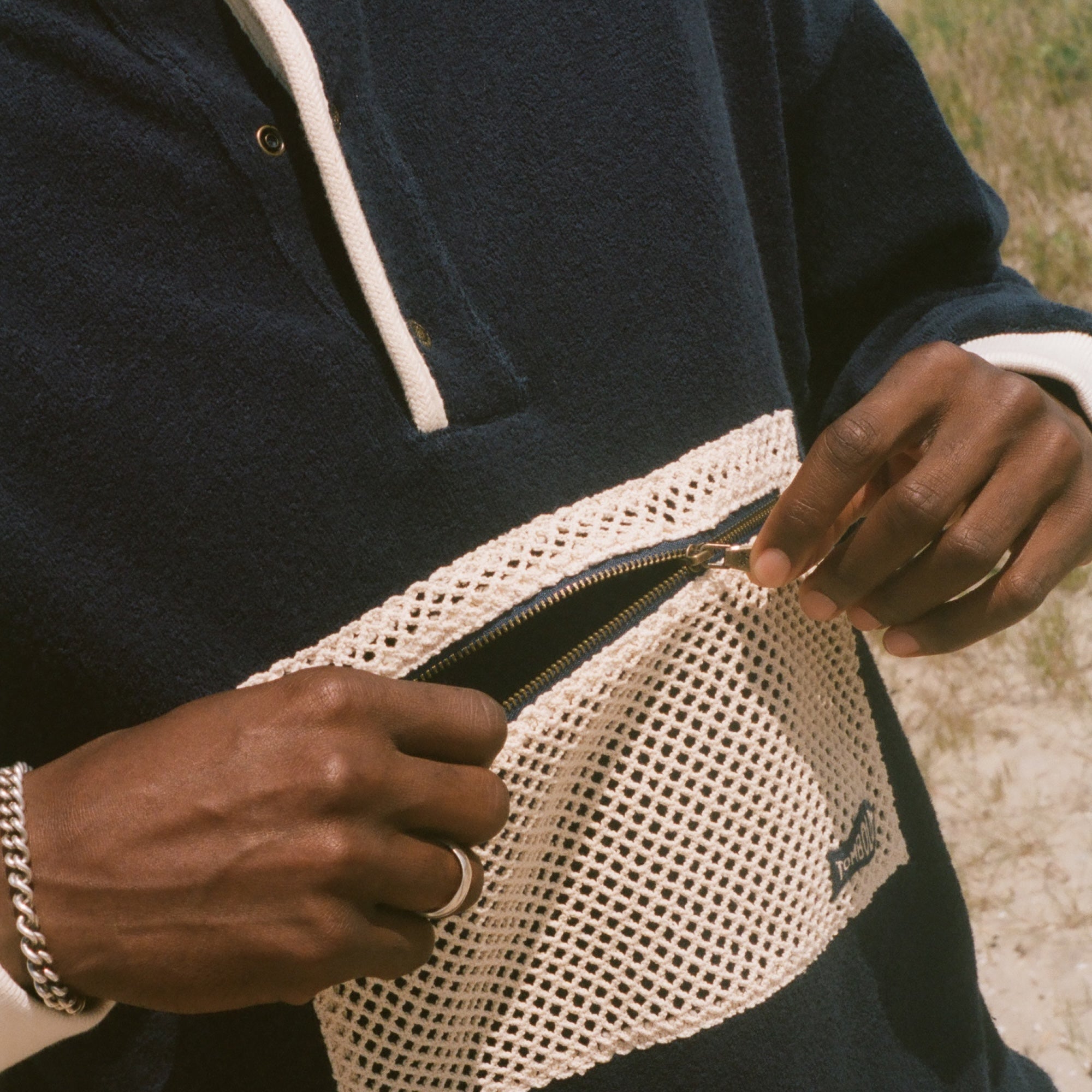 A close-up of a person outdoors wearing a navy long-sleeve pullover with white contrast trim and a large white mesh front pocket, with the zipper being opened. Sand and ocean are visible in the background.