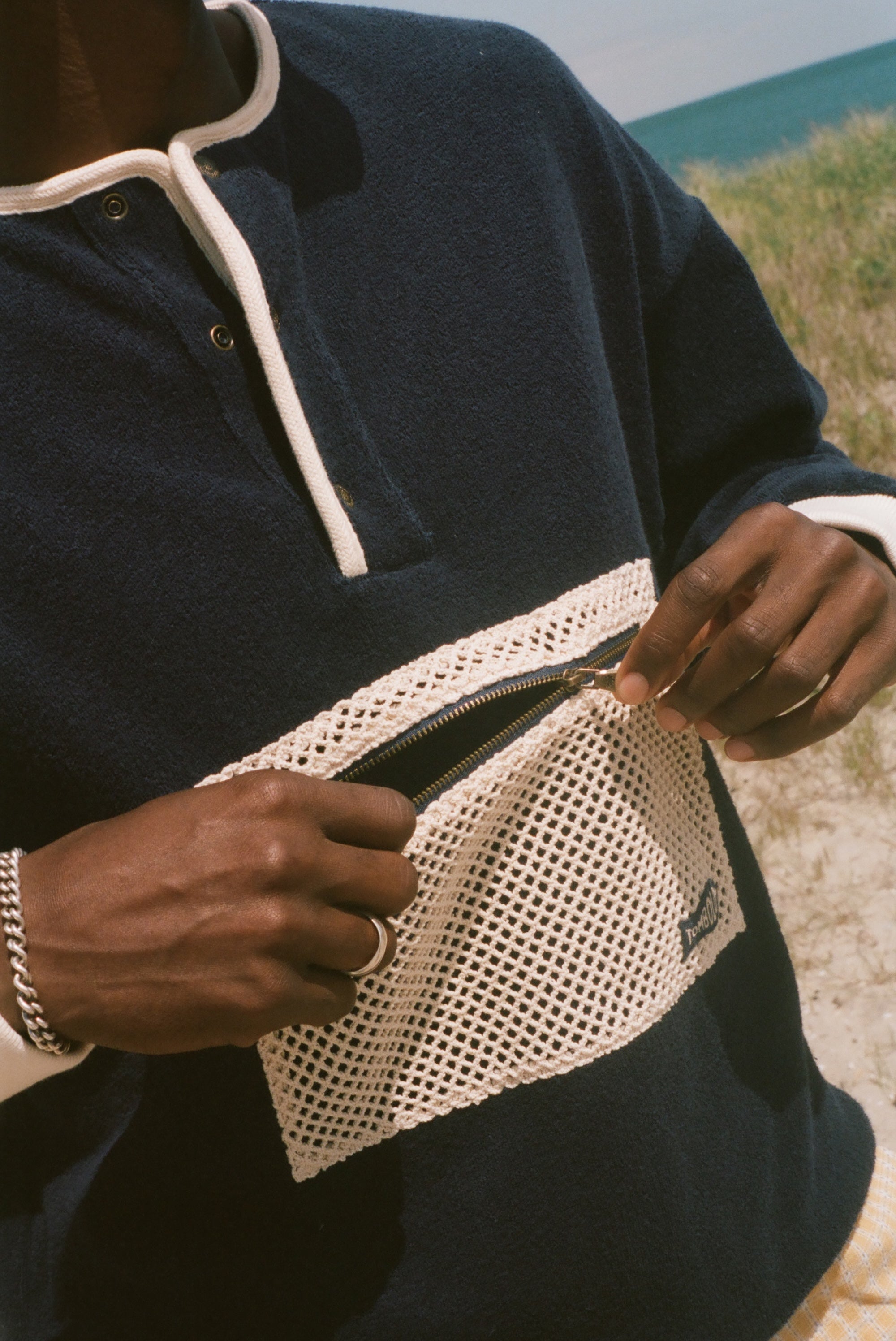 A close-up of a person outdoors wearing a navy long-sleeve pullover with white contrast trim and a large white mesh front pocket, with the zipper being opened. Sand and ocean are visible in the background.