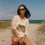 Woman in a yellow t-shirt with 'Turismo' printed on it, sitting on a sandy beach with ocean and blue sky in the background.