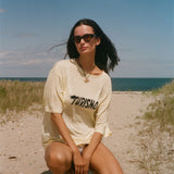 Woman in a yellow t-shirt with 'Turismo' printed on it, sitting on a sandy beach with ocean and blue sky in the background.
