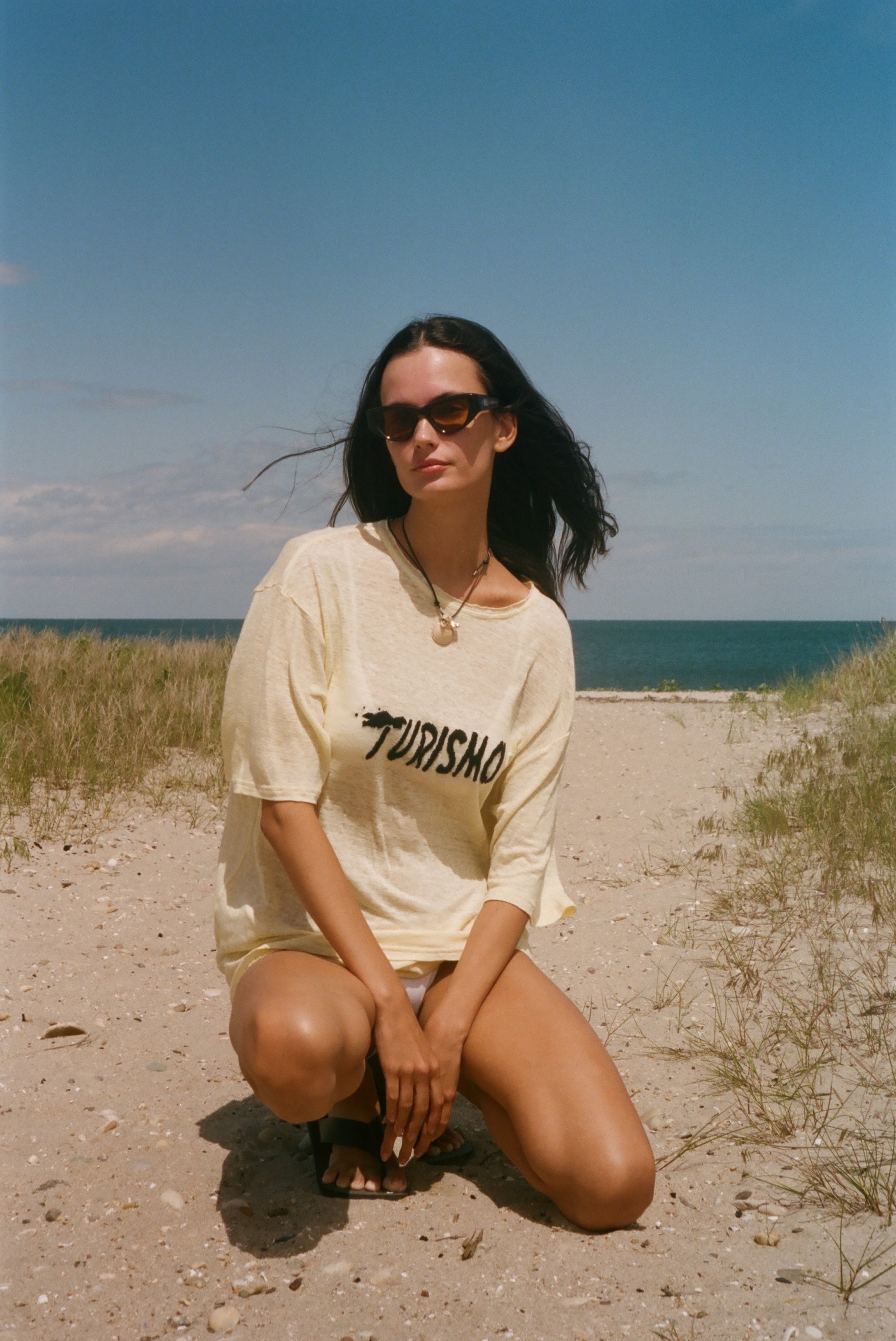 Woman in a yellow t-shirt with 'Turismo' printed on it, sitting on a sandy beach with ocean and blue sky in the background.