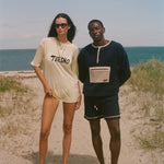 Two people standing on a sandy beach with ocean in the background. The woman is wearing an oversized 'Turismo' t-shirt.
