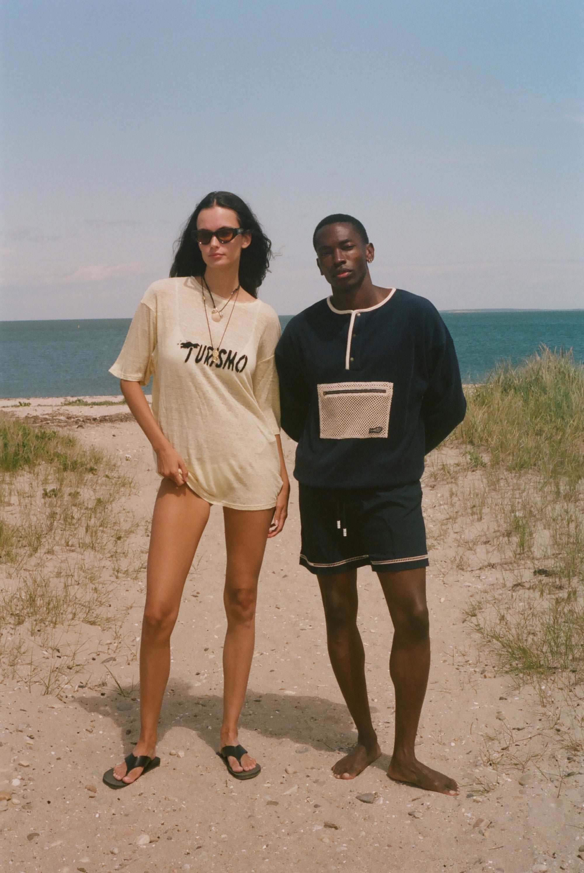 Two people standing on a sandy beach with ocean in the background. The woman is wearing an oversized 'Turismo' t-shirt.