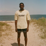 Person standing on a sandy beach with ocean in the background, wearing a yellow linen 'Turismo' tee.