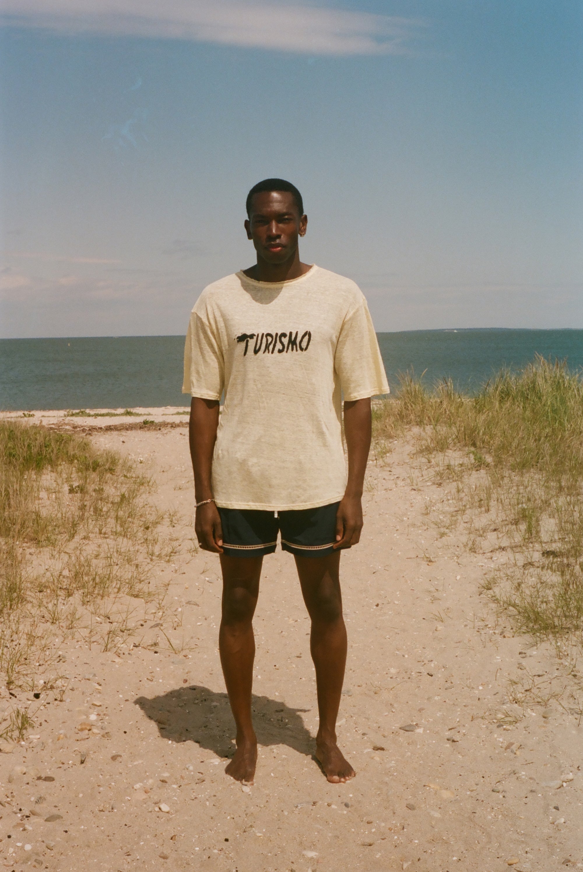 Person standing on a sandy beach with ocean in the background, wearing a yellow linen 'Turismo' tee.
