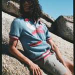 Male model with long black hair wearing blue cabana shirt with lobster while leaning on a rock wearing gray shorts 