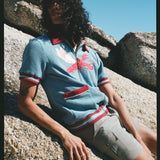 Male model with long black hair wearing blue cabana shirt with lobster while leaning on a rock wearing gray shorts 