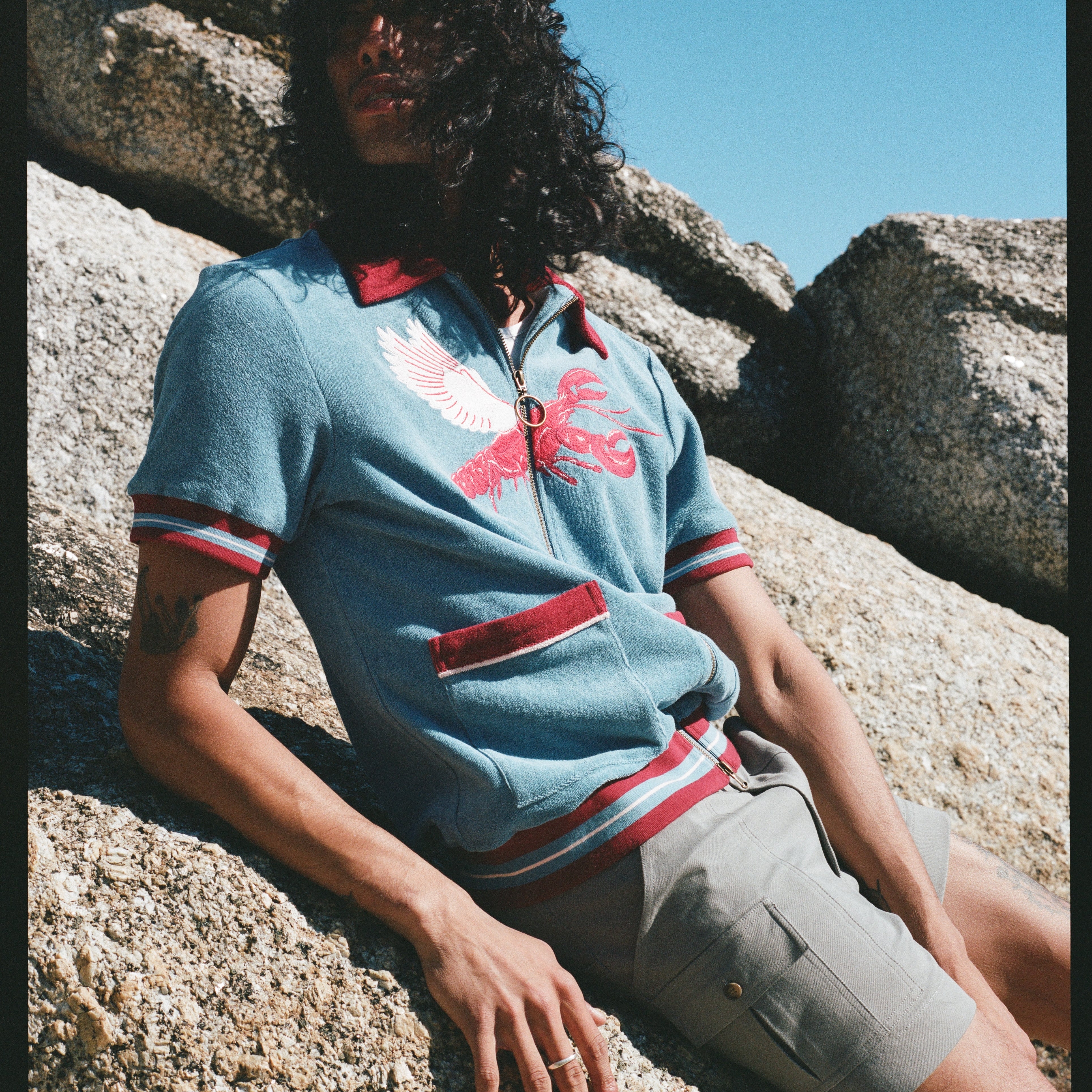 Male model with long black hair wearing blue cabana shirt with lobster while leaning on a rock wearing gray shorts 