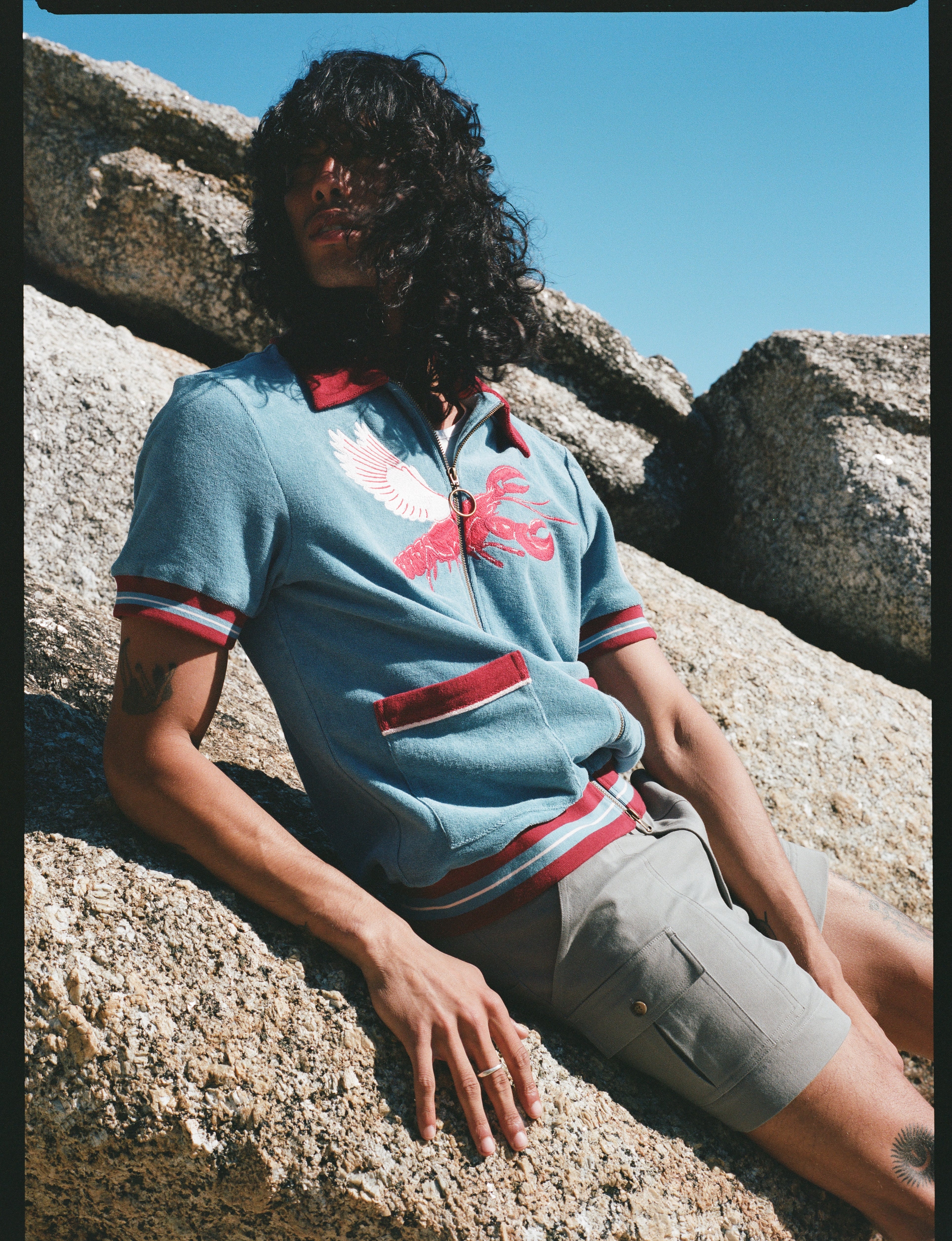 Male model with long black hair wearing blue cabana shirt with lobster while leaning on a rock wearing gray shorts 