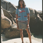 Male model with long black hair wearing blue cabana shirt with lobster standing in front of large boulders wearing gray shorts