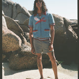 Male model with long black hair wearing blue cabana shirt with lobster standing in front of large boulders wearing gray shorts