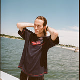 Male model with shoulder length brown hair running his hands through his hair while wearing the 'Boston Whaler' terry tee, shot on a background of the water, a thin strip of land, and blue skies.