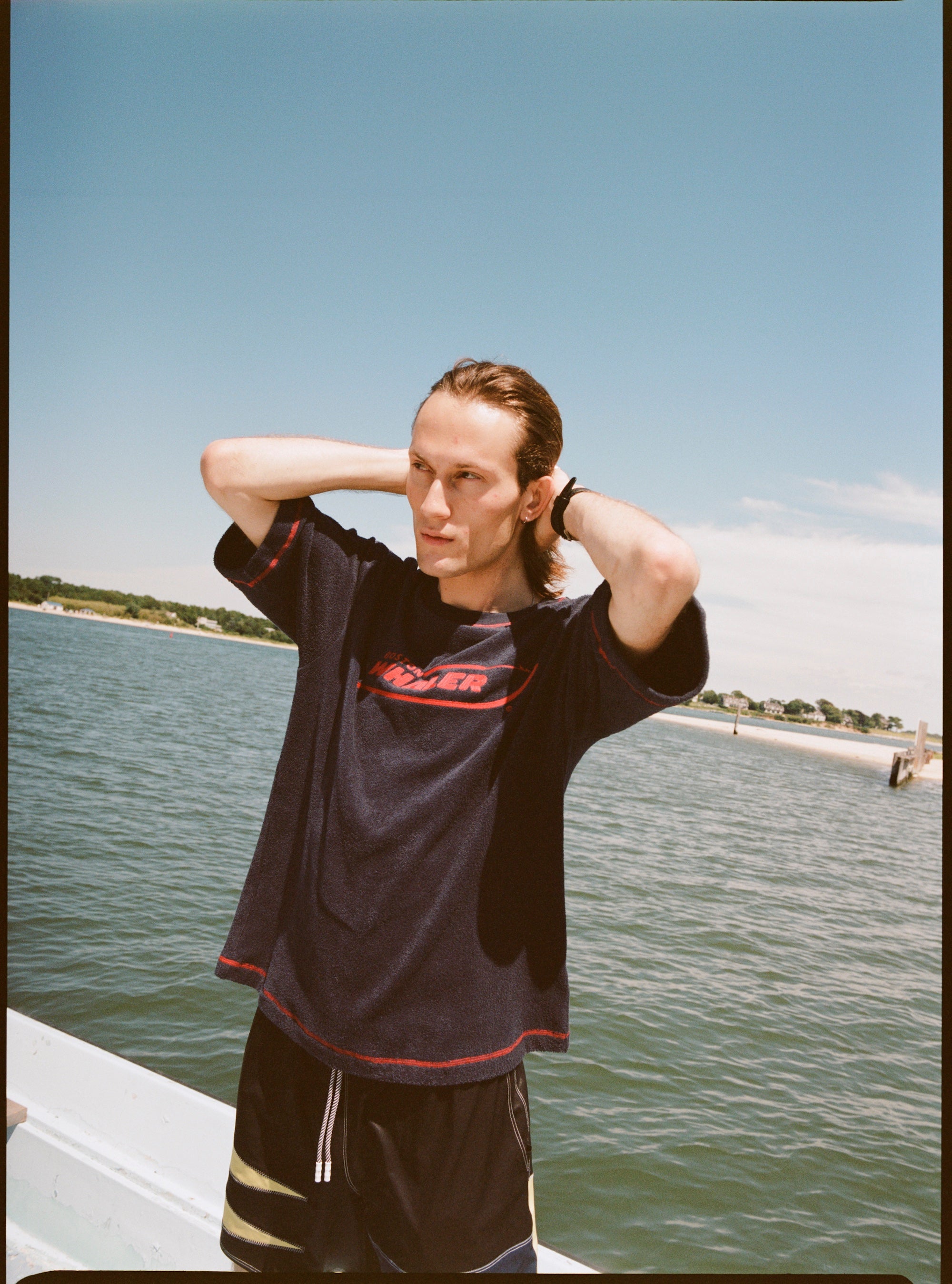 Male model with shoulder length brown hair running his hands through his hair while wearing the 'Boston Whaler' terry tee, shot on a background of the water, a thin strip of land, and blue skies.