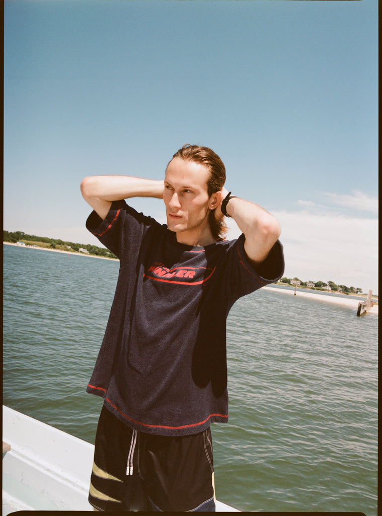 Male model with shoulder length brown hair running his hands through his hair while wearing the 'Boston Whaler' terry tee, shot on a background of the water, a thin strip of land, and blue skies.