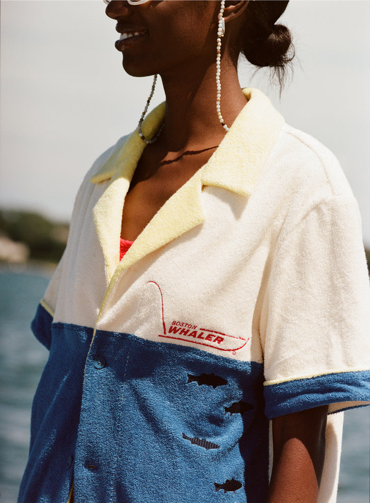 Female model wearing white sunglasses and a 'boston whaler' no nibbles cabana with a red swimsuit underneath, shot on a background of water, a thin strip of land, and blue skies.