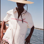 Female model wearing a large beige sun hat and a 'Boston Whaler' Cabana (white), shot on a background of water, a thin strip of land, and blue skies.