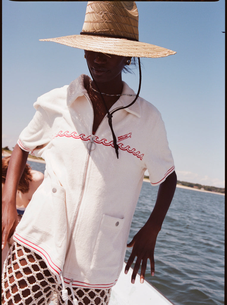 Female model wearing a large beige sun hat and a 'Boston Whaler' Cabana (white), shot on a background of water, a thin strip of land, and blue skies.