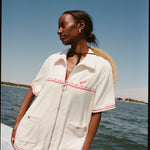 Female model with a sunhat hanging off her back while wearing the 'Boston Whaler' Cabana (white), shot on a background of the water, shot on a thin strip of land, and blue skies.