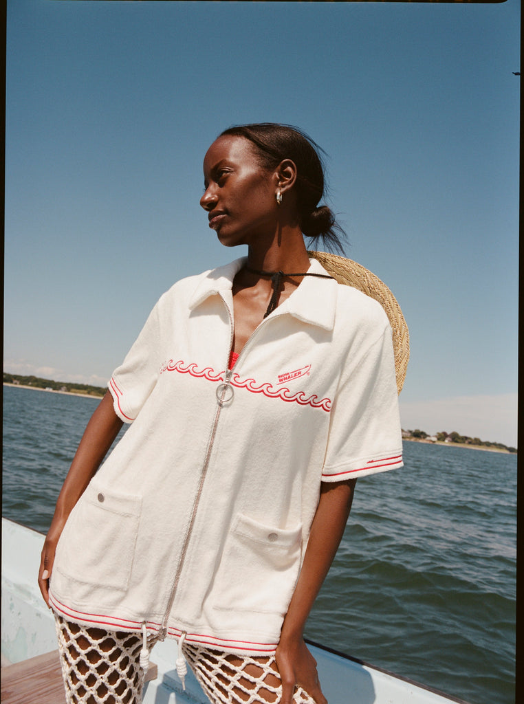 Female model with a sunhat hanging off her back while wearing the 'Boston Whaler' Cabana (white), shot on a background of the water, shot on a thin strip of land, and blue skies.