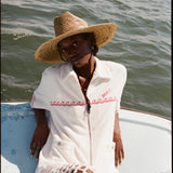Female model wearing a large beige sunhat, the 'Boston Whaler' cabana in white, white netting pants, shot on a background of the water.