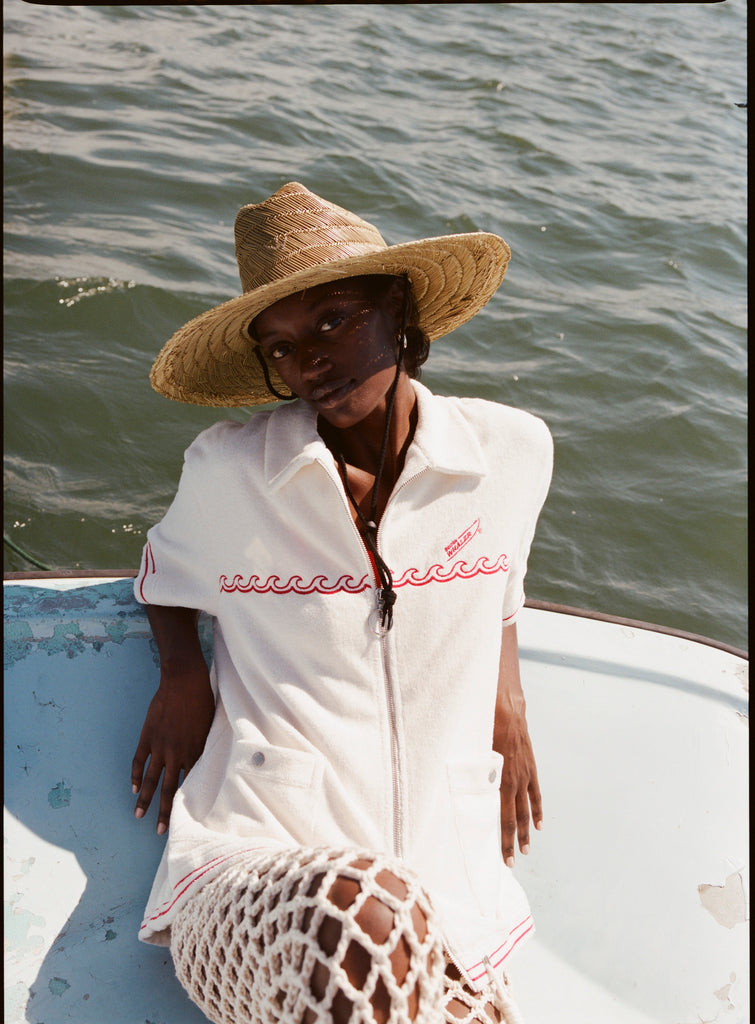 Female model wearing a large beige sunhat, the 'Boston Whaler' cabana in white, white netting pants, shot on a background of the water.