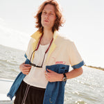 Male model wearing with shoulder length brown hair standing on a boat wearing the 'boston whaler' no nibbles cabana unbuttoned with a white shirt underneath and black swim trunks with a pair sunglasses hanging from his neck.