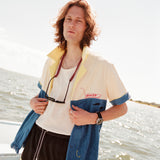 Male model wearing with shoulder length brown hair standing on a boat wearing the 'boston whaler' no nibbles cabana unbuttoned with a white shirt underneath and black swim trunks with a pair sunglasses hanging from his neck.