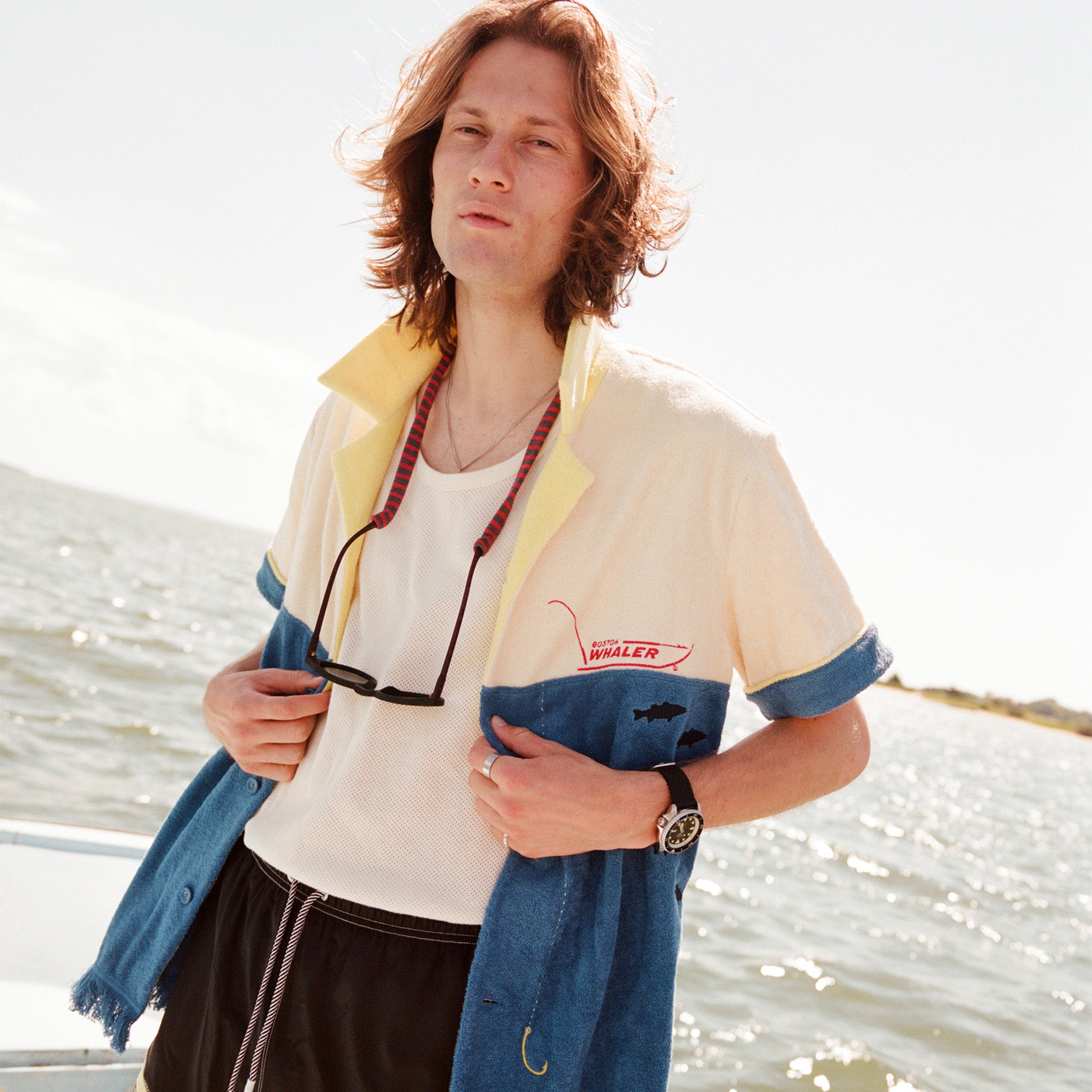 Male model wearing with shoulder length brown hair standing on a boat wearing the 'boston whaler' no nibbles cabana unbuttoned with a white shirt underneath and black swim trunks with a pair sunglasses hanging from his neck.