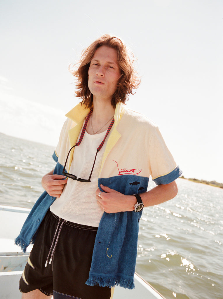 Male model wearing with shoulder length brown hair standing on a boat wearing the 'boston whaler' no nibbles cabana unbuttoned with a white shirt underneath and black swim trunks with a pair sunglasses hanging from his neck.
