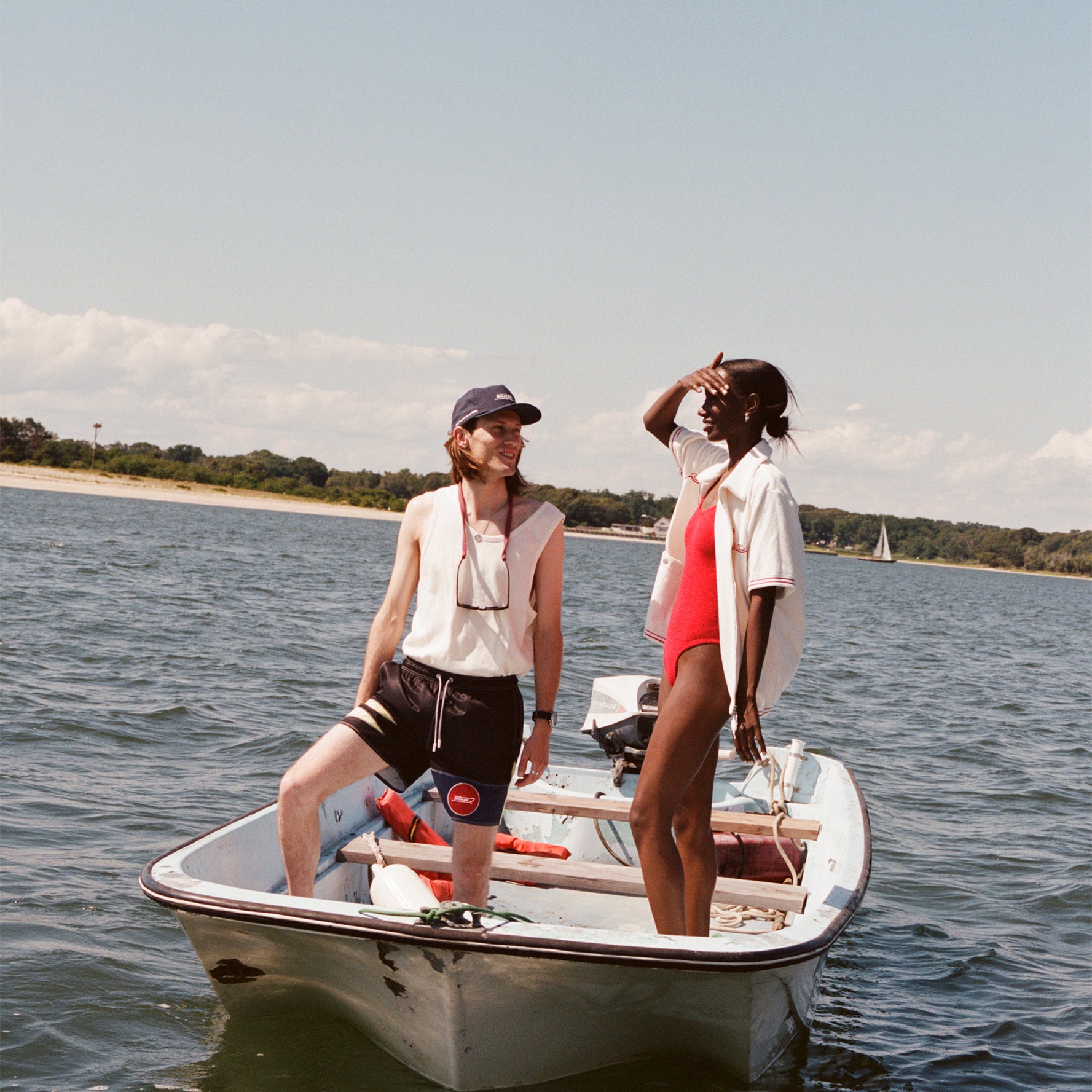 Male model with shoulder length brown hair wearing the 'boston whaler' swim trunks, a white tank top, and navy boston whaler souvenir cap, standin alognside a female model with black hair in a bun wearing a red swimswuit and white boston whaler cabana, shot on a boston whaler on the water with a strip of land and blue skies visible.