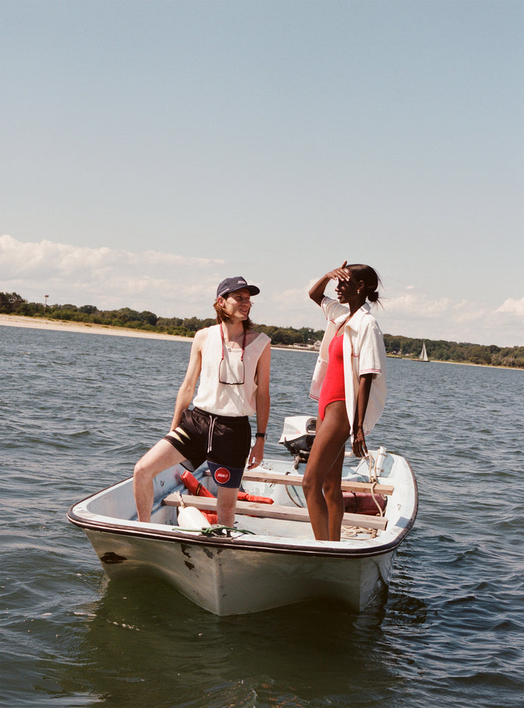 Male model with shoulder length brown hair wearing the 'boston whaler' swim trunks, a white tank top, and navy boston whaler souvenir cap, standin alognside a female model with black hair in a bun wearing a red swimswuit and white boston whaler cabana, shot on a boston whaler on the water with a strip of land and blue skies visible.