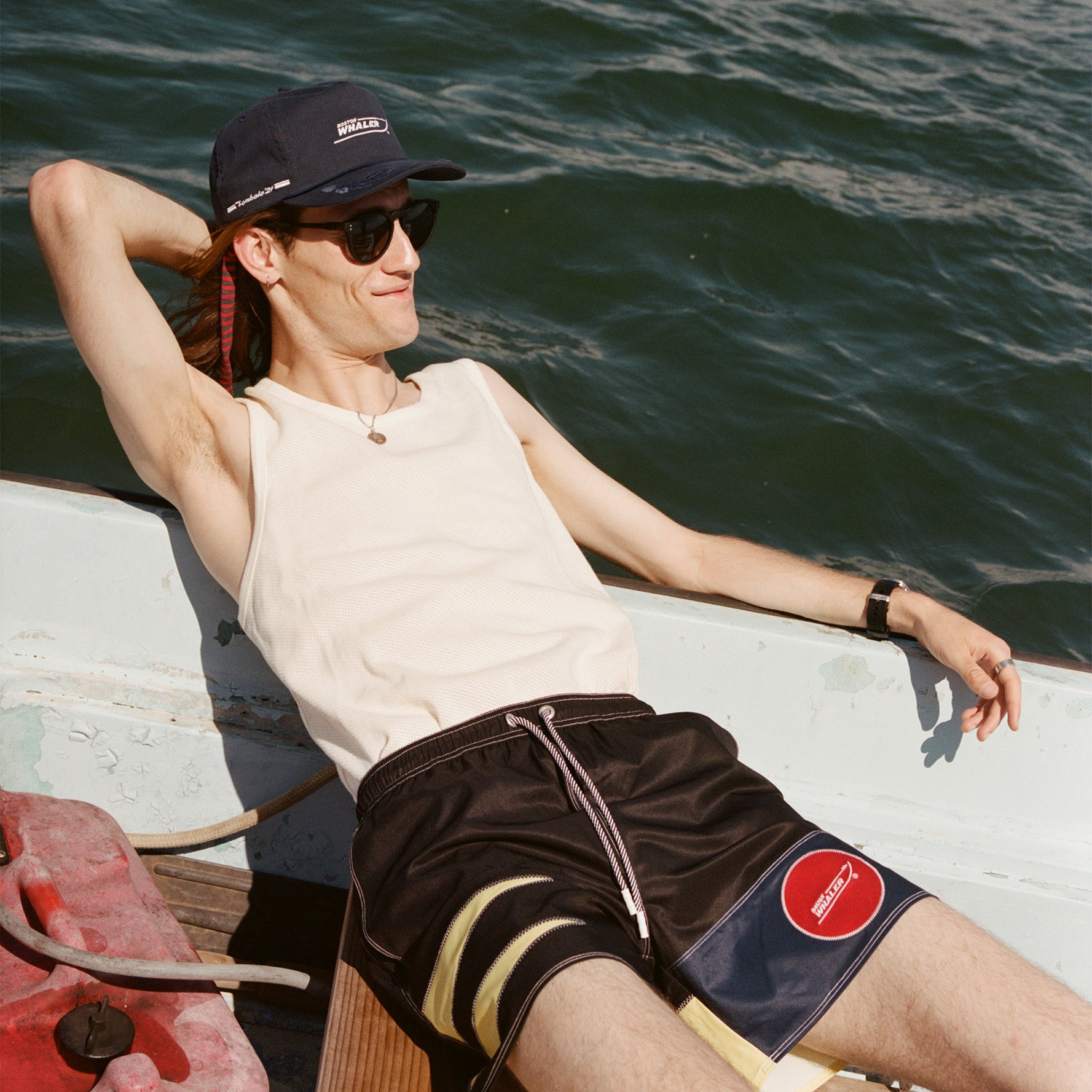 Male model with long brown hair wearing the 'Boston Whaler' 5-Panel Souvenir Cap while leaning back off the bow of a boat with one hand behind his head and the other resting on the boat.