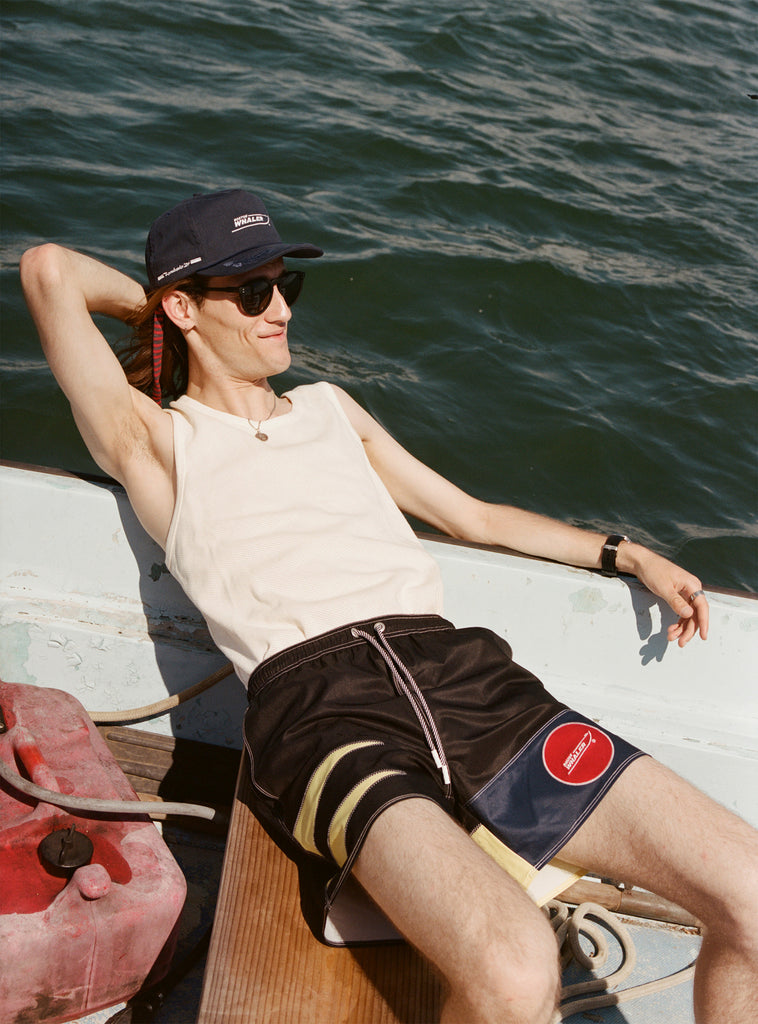 Male model with long brown hair wearing the 'Boston Whaler' 5-Panel Souvenir Cap while leaning back off the bow of a boat with one hand behind his head and the other resting on the boat.