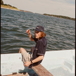 Male model wearing the 'boston whaler' cabana (navy) with a matching 'boston whaler' souvenir cap, and khaki shorts, sitting with his legs of the side of the boat, shot on a background of the water, a thin strip of land, and blue skies.