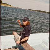Male model wearing the 'boston whaler' cabana (navy) with a matching 'boston whaler' souvenir cap, and khaki shorts, sitting with his legs of the side of the boat, shot on a background of the water, a thin strip of land, and blue skies.