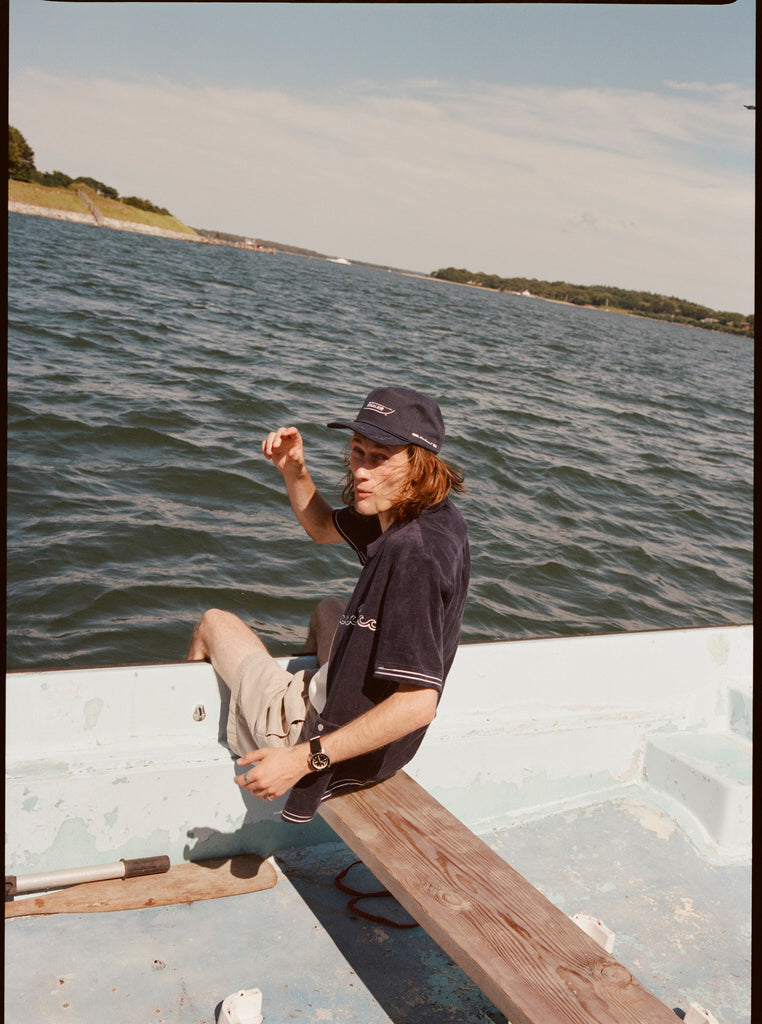 Male model wearing the 'boston whaler' cabana (navy) with a matching 'boston whaler' souvenir cap, and khaki shorts, sitting with his legs of the side of the boat, shot on a background of the water, a thin strip of land, and blue skies.