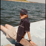 Male model wearing the 'boston whaler' cabana (navy) with a matching 'boston whaler' souvenir cap, and khaki shorts, sitting with his legs of the side of the boat, shot on a background of the water, a thin strip of land, and blue skies.