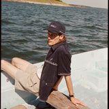Male model wearing the 'boston whaler' cabana (navy) with a matching 'boston whaler' souvenir cap, and khaki shorts, sitting with his legs of the side of the boat, shot on a background of the water, a thin strip of land, and blue skies.