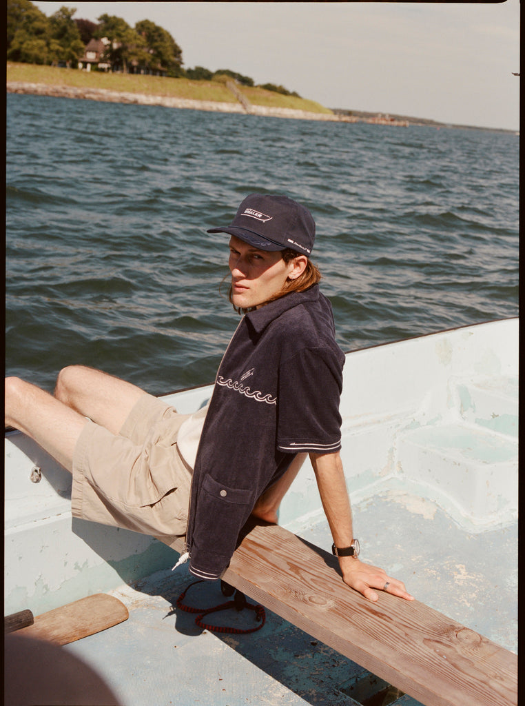 Male model wearing the 'boston whaler' cabana (navy) with a matching 'boston whaler' souvenir cap, and khaki shorts, sitting with his legs of the side of the boat, shot on a background of the water, a thin strip of land, and blue skies.
