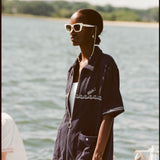 Female model with white sunglasses wearing the 'Boston Whaler' Cabana (Navy) half-zipped, standing on a boat, shot on a background of the water, a thin strip of land, and blue skies.