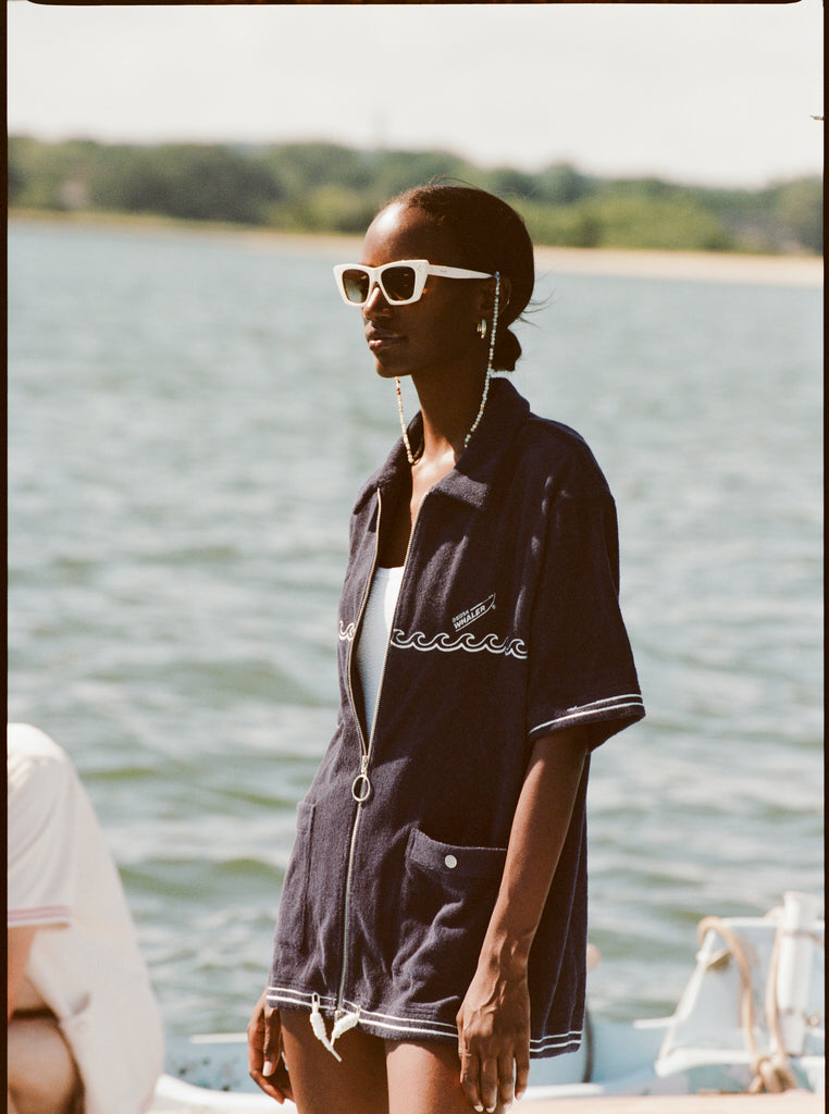 Female model with white sunglasses wearing the 'Boston Whaler' Cabana (Navy) half-zipped, standing on a boat, shot on a background of the water, a thin strip of land, and blue skies.