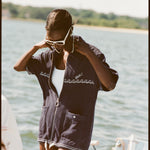 Female model with white sunglasses wearing the 'Boston Whaler' Cabana (navy) half-zipped while standing on a boat, with a background of the water, a thin strip of land, and blue skies.