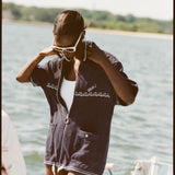 Female model with white sunglasses wearing the 'Boston Whaler' Cabana (navy) half-zipped while standing on a boat, with a background of the water, a thin strip of land, and blue skies.