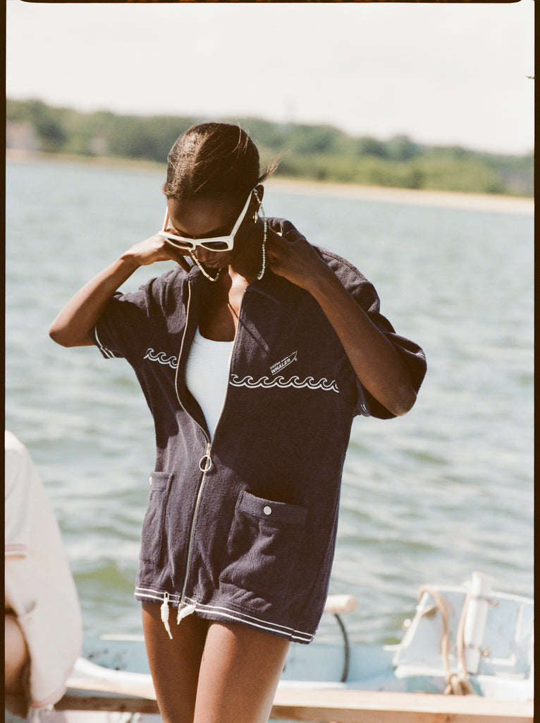 Female model with white sunglasses wearing the 'Boston Whaler' Cabana (navy) half-zipped while standing on a boat, with a background of the water, a thin strip of land, and blue skies.