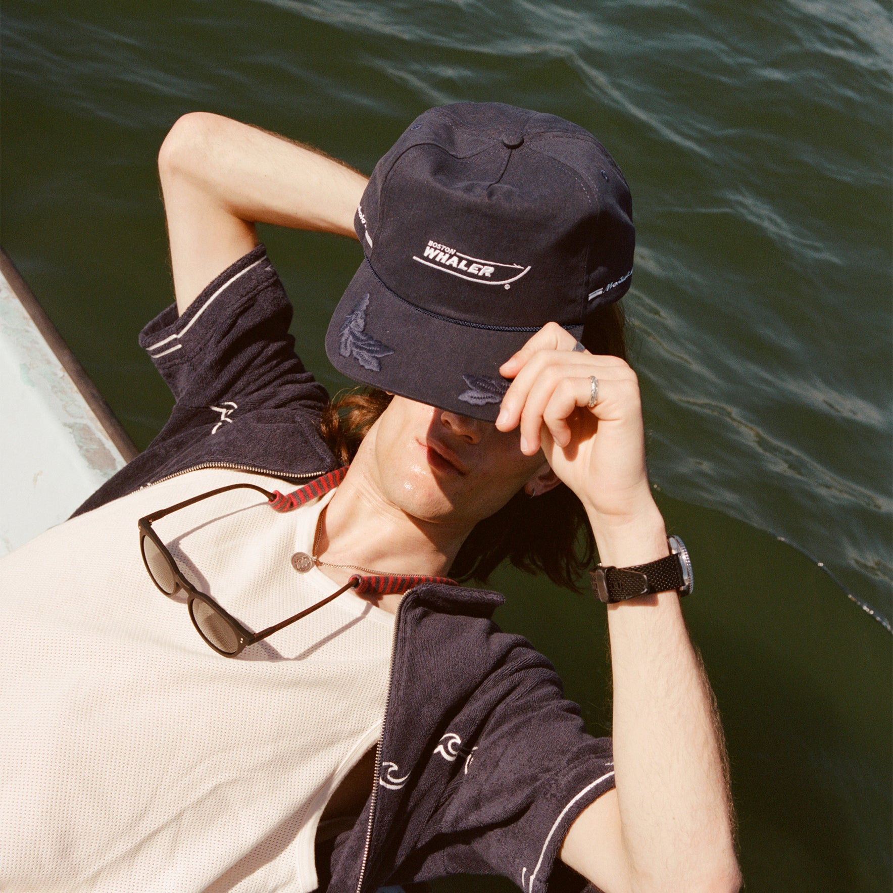 Male model with long brown hair lifting the 'Boston Whaler' 5-Panel Souvenir Cap off his head while leaning off the side of a boat.