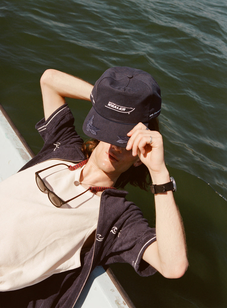 Male model with long brown hair lifting the 'Boston Whaler' 5-Panel Souvenir Cap off his head while leaning off the side of a boat.