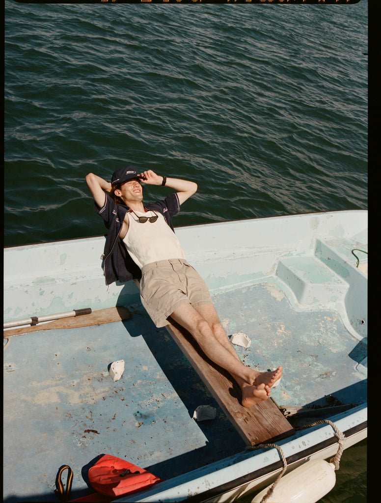 Male model wearing the 'Boston Whaler' Cabana (navy) while lying on a boat with his head over the bow, and one arm supporting it, shot on a background of the water.