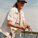 Male model with shoulder length brown hair wearing the 'Boston Whaler' 5-Panel Souvenir Cap while holding a line on a boat, shot on a background of the water, a thin strip of land, and blue skies.