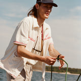 Male model with shoulder length brown hair wearing the 'Boston Whaler' 5-Panel Souvenir Cap while holding a line on a boat, shot on a background of the water, a thin strip of land, and blue skies.